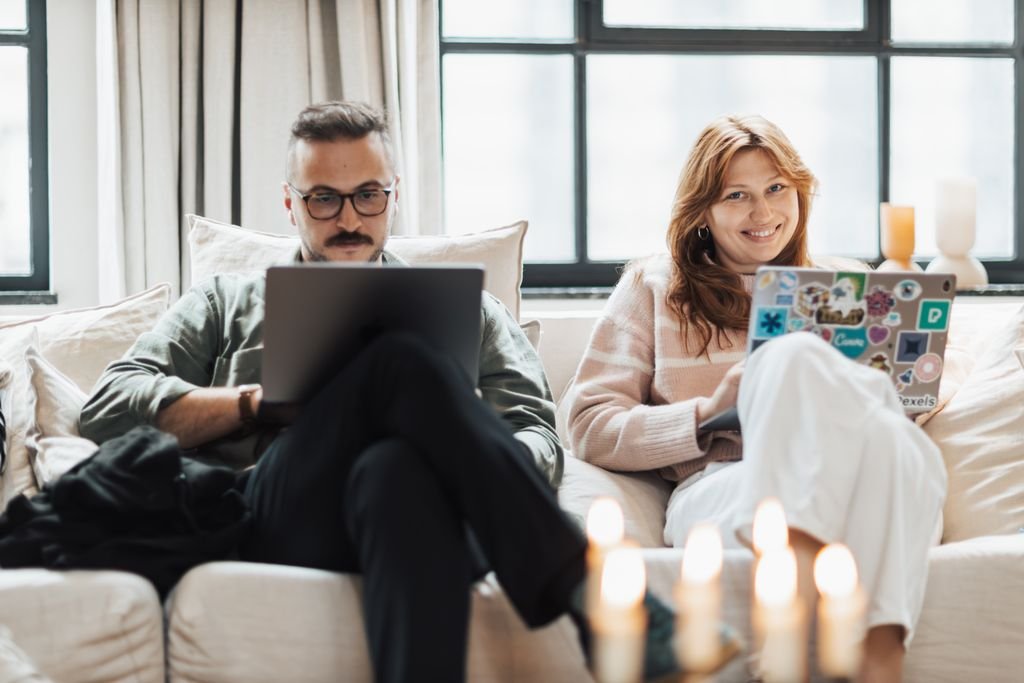 A man and woman work with laptops on a couch, enjoying a cozy indoor setting.